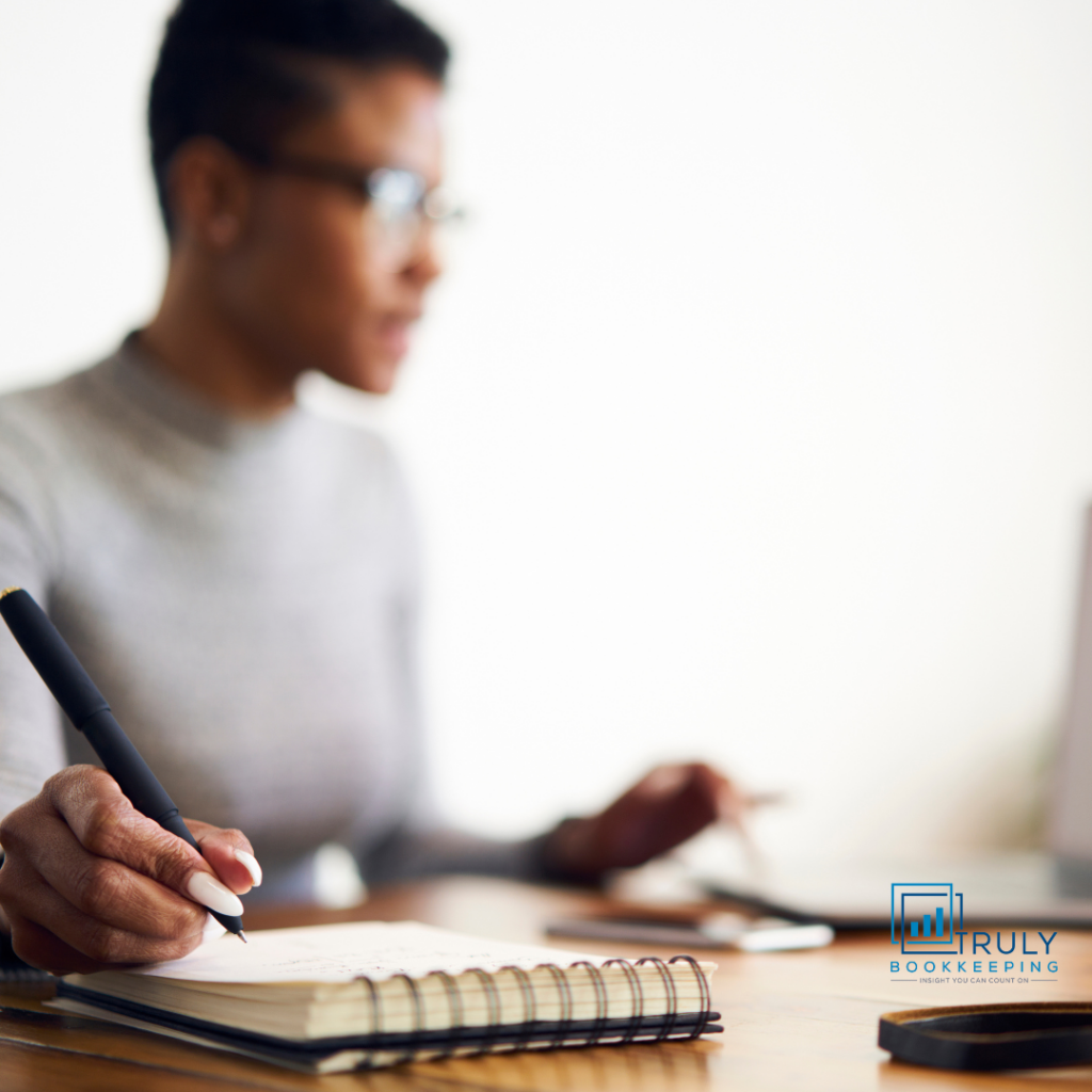 A professional woman sitting at a desk, focused on taking notes in a spiral notebook while working on her laptop. Truly Bookkeeping logo in the bottom right corner.