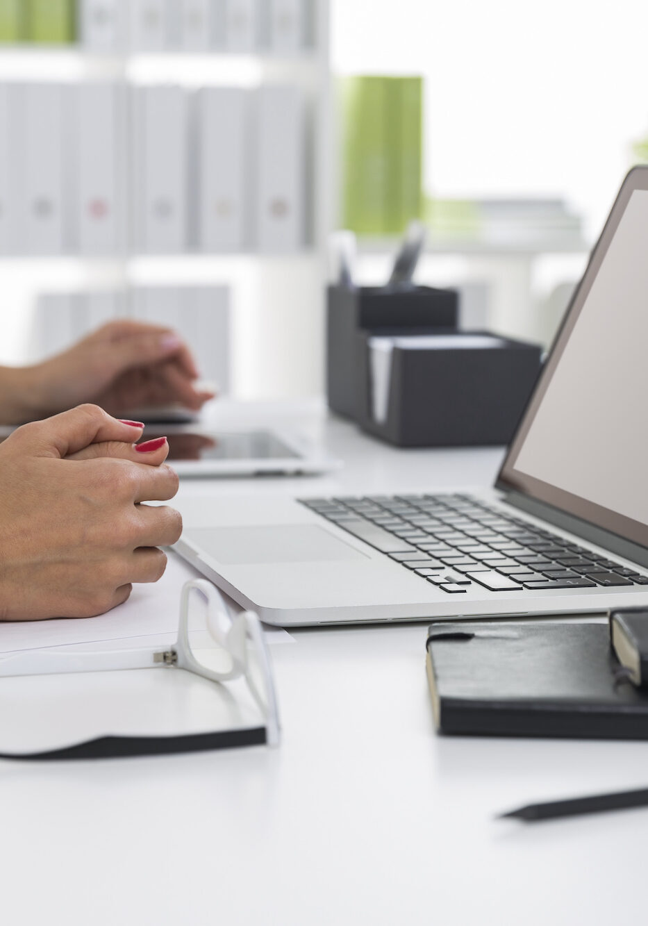 Close up of two pairs of women's hands in an office. Blank laptop screen. Glasses and notebooks are on the table.